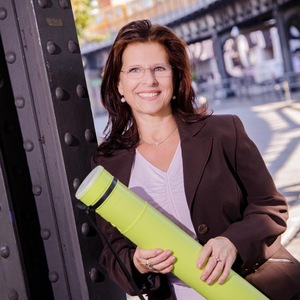 Portraitfoto einer Unternehmerin im Businessoutfit, stehend an einem Pfeiler der Hochbahn mit heranfahrender U-Bahn im Hintergrund