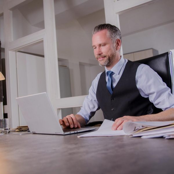 Corporate Fotografie in Berlin - Imagefoto eines Unternehmers im Businessoutfit bei der Arbeit in seinem Büro am Schreibtisch sitzend und auf sein aufgeklapptes Laptop blickend