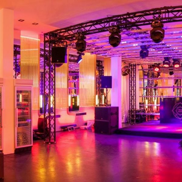 Interieurfoto eines mit Effektlichtern beleuchteten Beautysalons, in dem sich zentral ein Lichtkäfig mit DJ Pult befindet