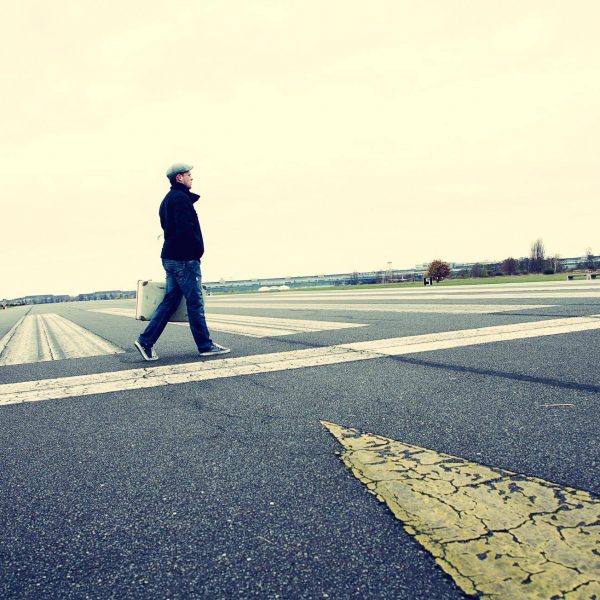 Imagefoto eines alten Reisekoffers, der von einer Person gehalten wird, welche die Startbahn des Tempelhofer Flugfelds überquert